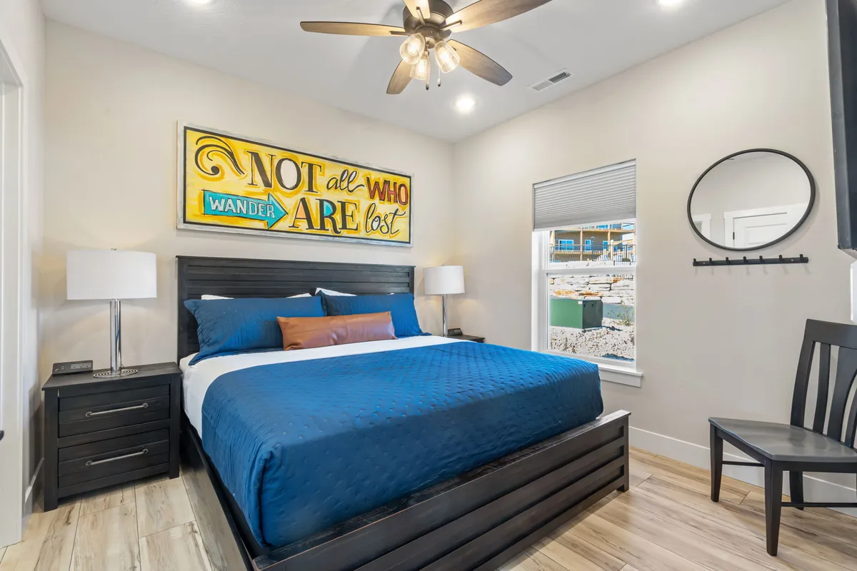 A tranquil bedroom featuring a plush bed with blue bedding, decorative wall art, a nightstand with a lamp, and a round mirror. The room is bathed in natural light, creating a relaxing and restful atmosphere.