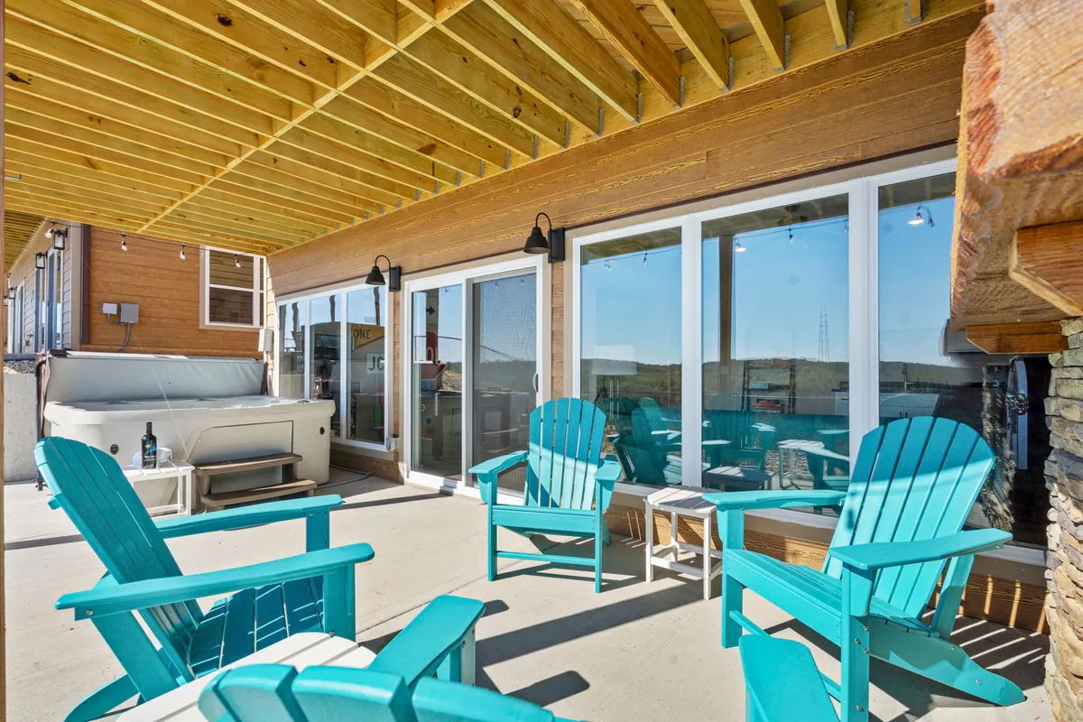 An exterior patio scene showing Adirondack chairs and a bubbling hot tub just outside large sliding doors. The wooden ceiling and scenic setting beckon for relaxation after indoor fun.