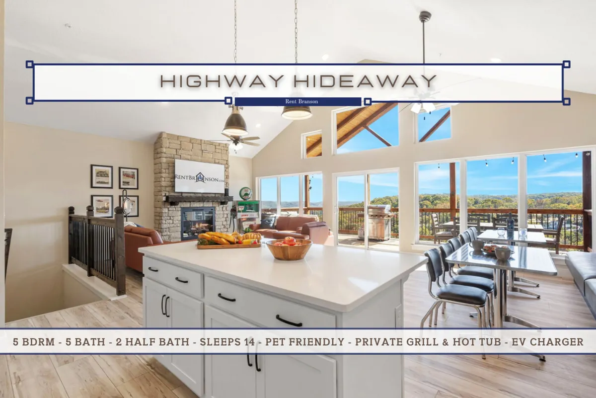 This image presents an expansive, open-concept living area featuring large windows that frame beautiful scenic views. The space flows seamlessly between a stone fireplace, a cozy living area with plush seating, a modern kitchen island, and a dining table, all bathed in natural light. The design highlights comfort and gathering, with direct visual access to the deck for indoor-outdoor living.