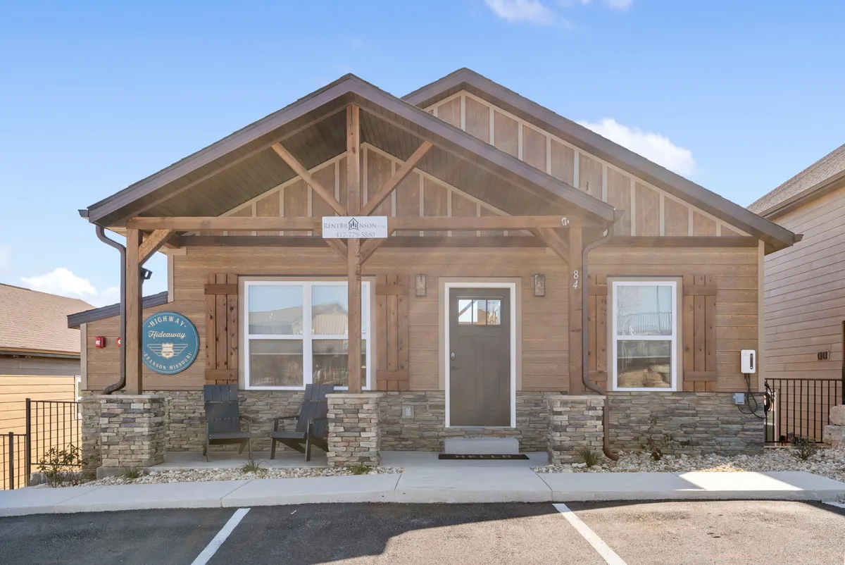 An exterior shot featuring a wooden structure with a stone facade, visible signage, and an inviting outdoor seating area. The porch area is set up for relaxation and socializing, with views that suggest easy access for travelers arriving by car.