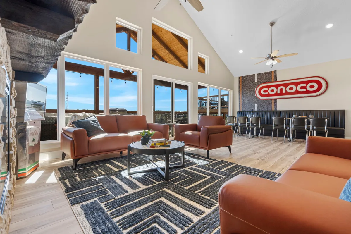 The image showcases a spacious and inviting living area with plush leather sofas centered around a stylish coffee table, all bathed in natural light from expansive windows that offer scenic views of the landscape. A vintage Conoco sign adds a nostalgic flair, while the adjacent dining area hints at memorable meals and social gatherings. The setup suggests a perfect place for guests to relax, socialize, and take in the tranquil surroundings.