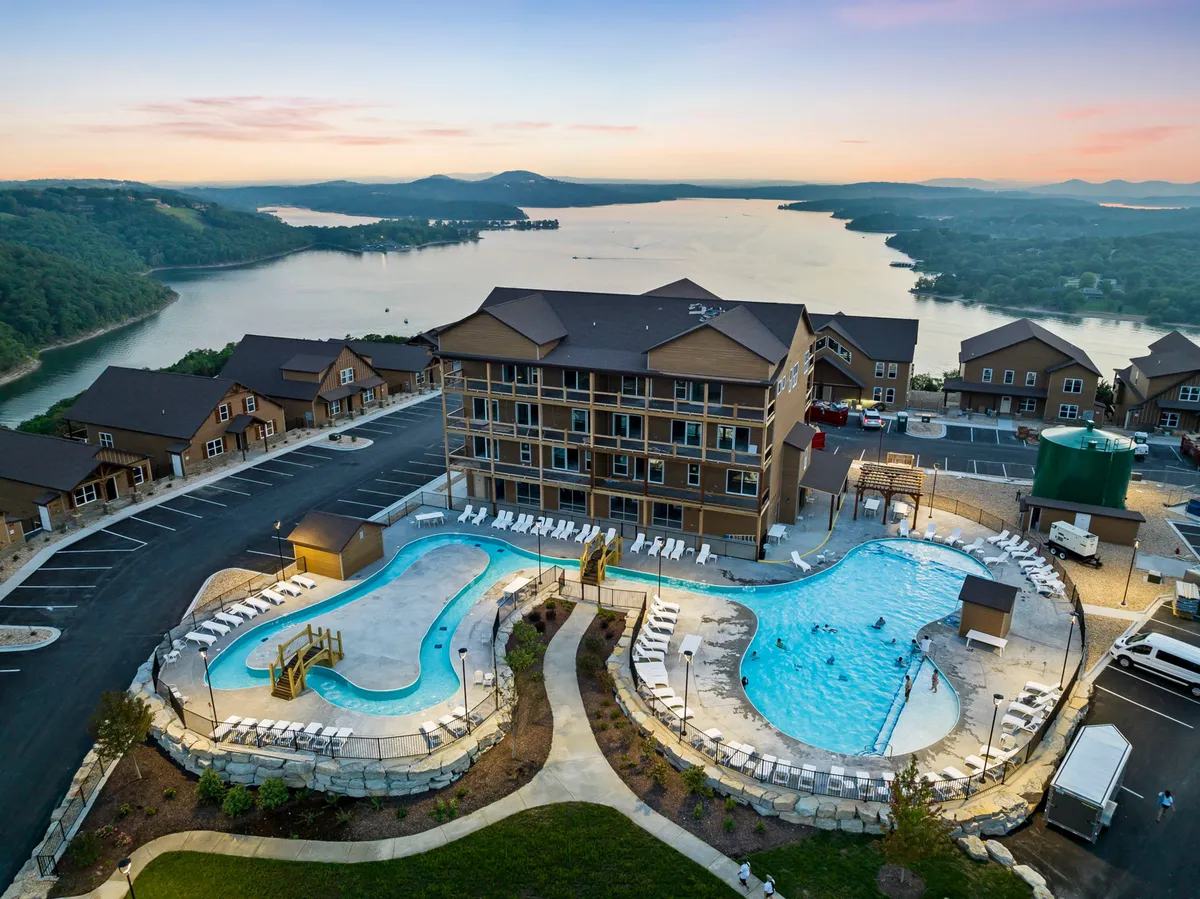 A cheerful exterior scene highlighting a resort-style swimming pool area with a clear view of the adjacent lake, the hotel building, and lush landscaping. Lounge chairs and the inviting water suggest a place for relaxation, social gatherings, and family fun. The setting sun casts a warm light over the community, capturing the vibrant and communal energy of the amenities.