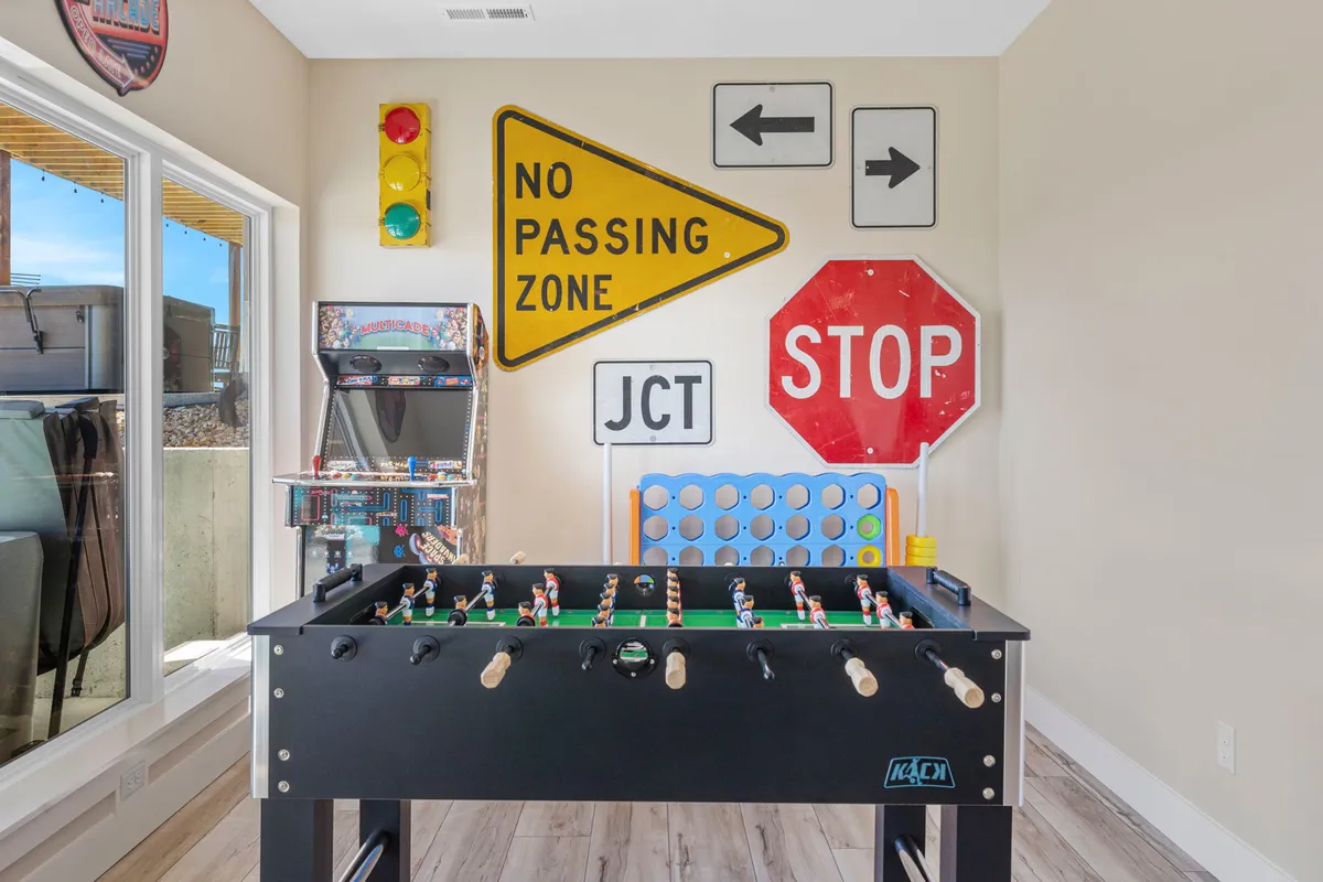 A vibrant game room featuring a prominent foosball table in the foreground, a classic arcade game machine against the wall, and playful traffic sign decor. The space is filled with energetic lighting, inviting guests to engage in friendly competitions and fun.