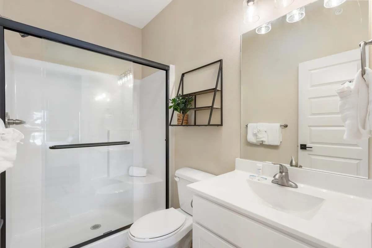 A modern ensuite bathroom featuring a glass shower enclosure, a sleek vanity with a mirror lit by soft lighting, and decorative shelving with plants. The atmosphere is fresh, spa-like, and inviting.