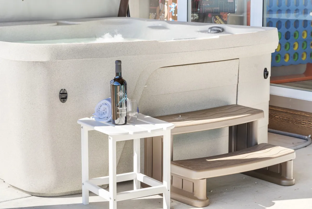 An inviting outdoor hot tub is set beside a small table holding a bottle of wine and a towel, offering a relaxing spot surrounded by privacy. Steps lead up to the tub, making it accessible for all guests.